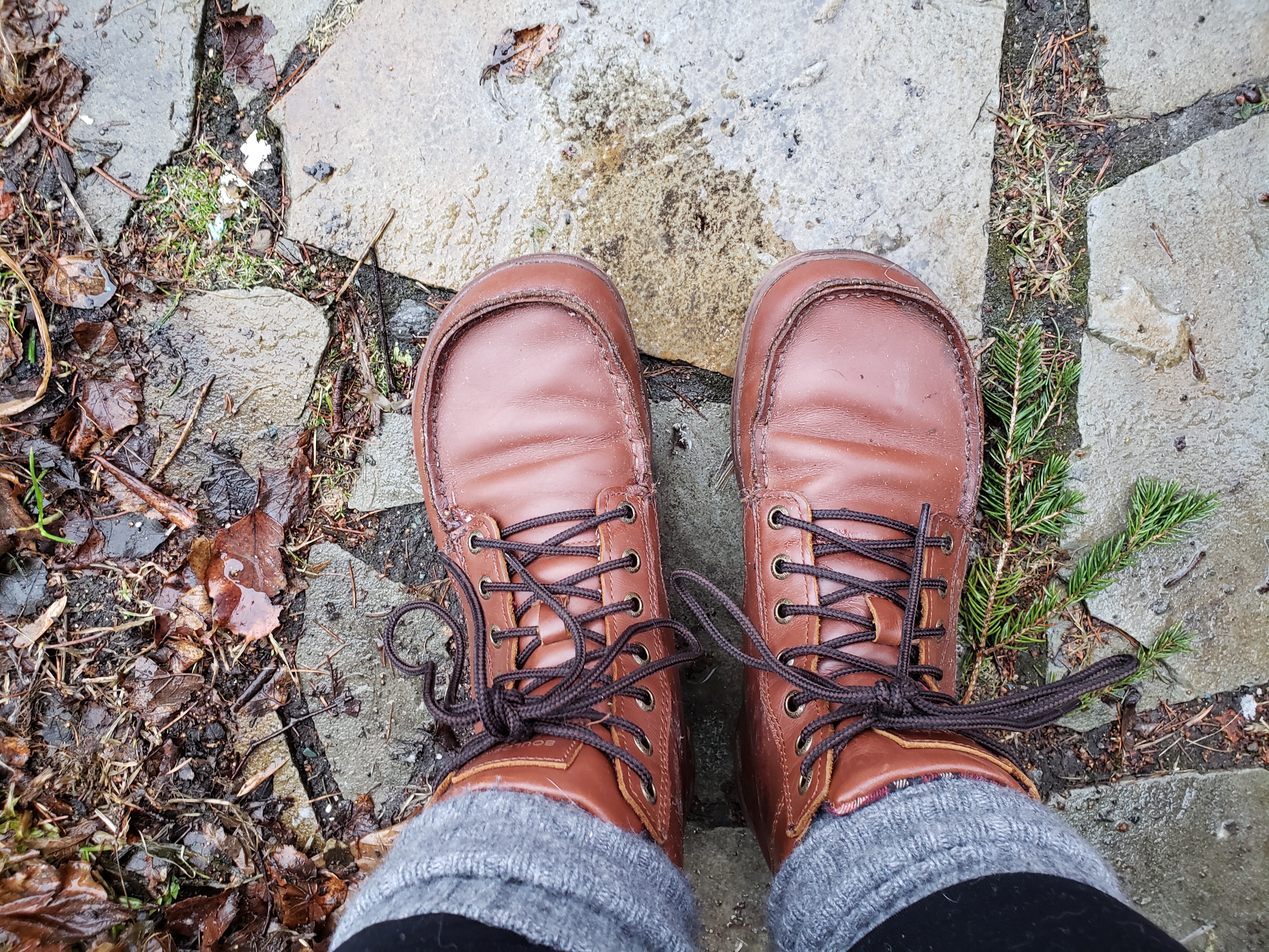 A person's hiking-boot-clad feet, shot from above. The boots are a russet-brown leather with dark brown laces. The person is also wearing grey wool socks and black leggings. They are standing on a rain-slicked flagstone path.