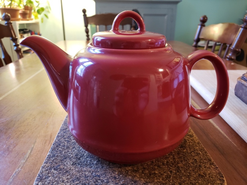 A cheerful, cherry-red teapot sits on a dark brown cork trivet on a wooden dining room table. To the right of the teapot is a cutting board, on which a chocolate babka is just visible. In the background are three chairs, a grey-blue wooden cabinet, and a shelf of houseplants in terra cotta pots.