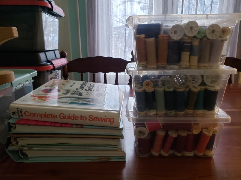 Newly organized sewing supplies on a dining room table. On the right are three stacked transparent plastic storage containers of thread; the bottom container is for reds, pinks, and purples, the middle one for blues and greens, and the top is for black, white, yellows, beiges and greys. To the left of the thread containers is a stack of four hardcover books, the topmost of which is the Reader's Digest Complete Guide to Sewing. To the left of the books sit three larger plastic bins.