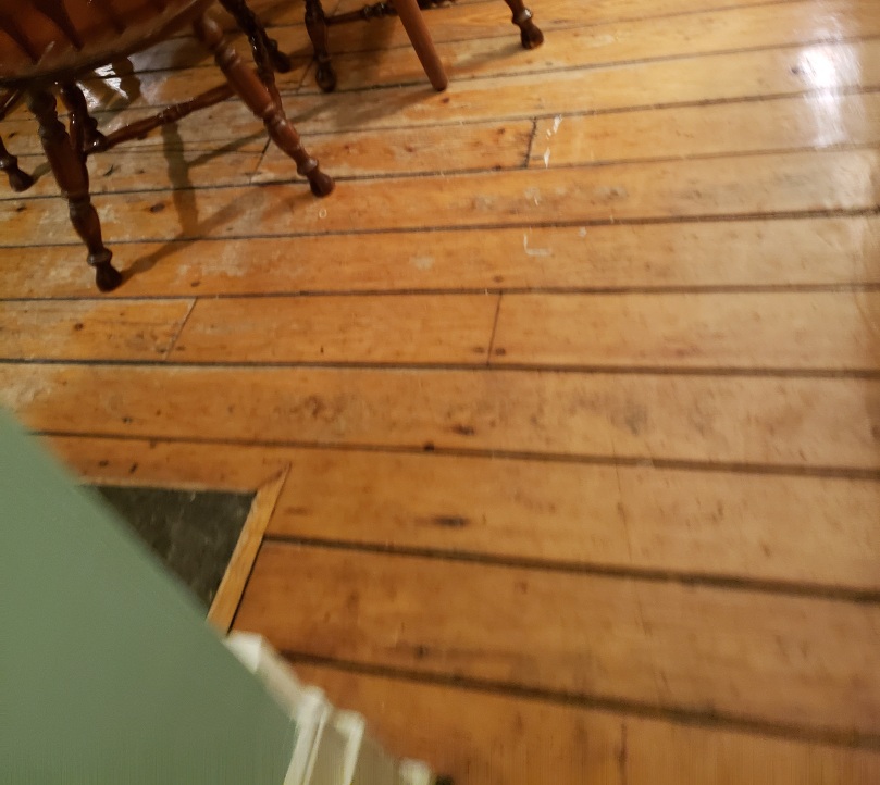 A slightly out-of-focus, weirdly angled shot of a softwood floor. The legs of three wooden country-style dining chairs are visible at the upper left corner. 